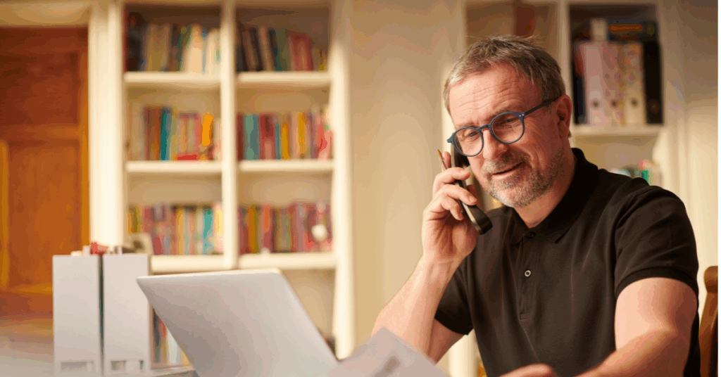 Middle-aged man talking on the phone while reviewing documents and using a laptop at home, representing financial planning for selling a business.